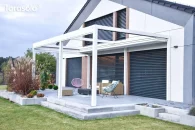 Pergola Tarasola Flat na 6 podporach w kolorze śnieżnej bieli tworzy nowoczesne, prostokątne zadaszenie nad tarasem z szarymi płytami. Ma tkaninowy dach i pełny układ podpór z każdej strony, a w tle widać ogród z ozdobnymi trawami i nowoczesną elewacją domu.