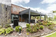 Pergola Elegancy Tarasola z elektrycznie zwijanym dachem i bocznymi roletami tworzy półzamkniętą, elegancką przestrzeń wypoczynkową z technorattanowymi meblami i huśtawką wiszącą. Wnętrze pergoli otwiera się na ogród z widokiem na ogród.