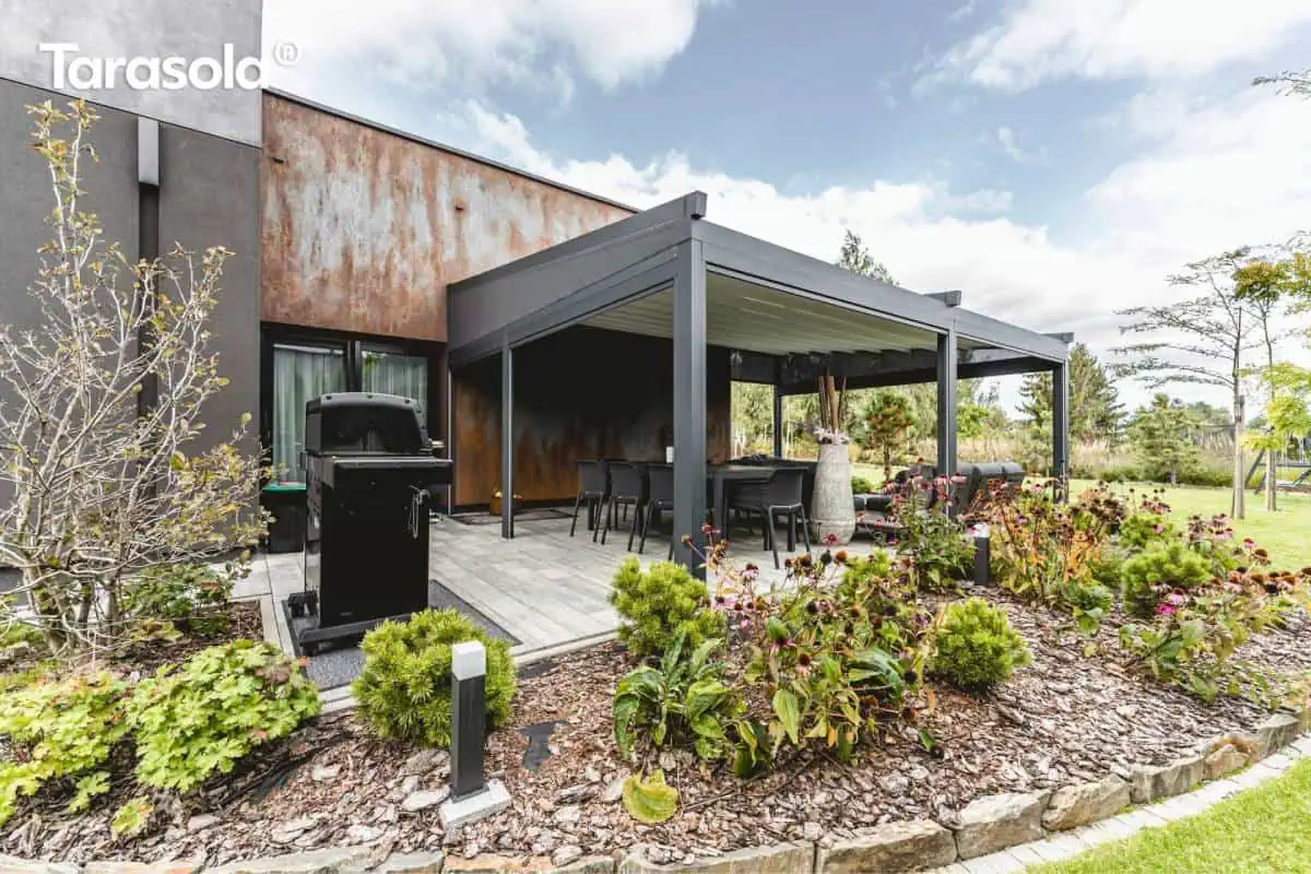 Taras zabudowany pergolą Tarasola Elegancy z roletami Pergola Elegancy Tarasola z elektrycznie zwijanym dachem i bocznymi roletami tworzy półzamkniętą, elegancką przestrzeń wypoczynkową z technorattanowymi meblami i huśtawką wiszącą. Wnętrze pergoli otwiera się na ogród z widokiem na ogród.