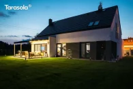 Pergola Elegancy Tarasola z ruchomym zadaszeniem tkaninowym tworzy nowoczesną, oświetloną strefę wypoczynkową na rozległym, zielonym trawniku. Pod pergolą ustawiono drewniany zestaw mebli, a otoczenie budynku podkreśla spokojny, wieczorny klimat.