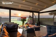 Pergola Elegancy Tarasola z równomiernie rozmieszczonym oświetleniem LED tworzy przytulne wnętrze tarasu wyposażonego w rattanowe sofy, stolik i dekoracje. Przestrzeń otwiera się na ogród z panoramicznym widokiem na pola i altanę ogrodową o zachodzie słońca.