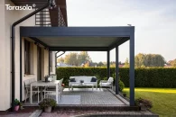 Nowoczesna pergola lamelowa Essential Tarasola zamocowana do ściany domu, prezentująca elegancką, antracytową konstrukcję z jasnymi lamelami dachowymi, które tworzą przyjemną, zacienioną przestrzeń wypoczynkową. Pod zadaszeniem ustawiono komfortowy zestaw mebli ogrodowych w jasnych barwach, a cały taras otacza perfekcyjnie przycięty, zielony żywopłot i zadbany trawnik, kreując spokojny i przytulny klimat.