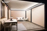 Pergola bioklimatyczna Essential z kompletem rolet bocznych ZIP, zapewniających prywatność i ochronę przed słońcem. Lamelowy dach w odcieniu kremowej bieli kontrastuje z antracytowymi profilami, tworząc elegancką strefę tarasową. Wnętrze tarasu urządzono minimalistycznymi meblami w jasnych kolorach, a otoczenie stanowi gęsty żywopłot oraz ogród pełen zieleni.