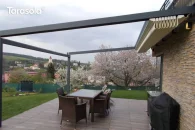 Pergola Elegancy Tarasola z całkowicie zwiniętym dachem rzymskim ułożonym w pakiet, odsłania aluminiową konstrukcję przyścienną nad tarasem z kompletem rattanowych mebli. Pod pergolą znajduje się przestrzeń jadalniana, otoczona ogrodem z kwitnącymi drzewami i widokiem na pobliską zabudowę.