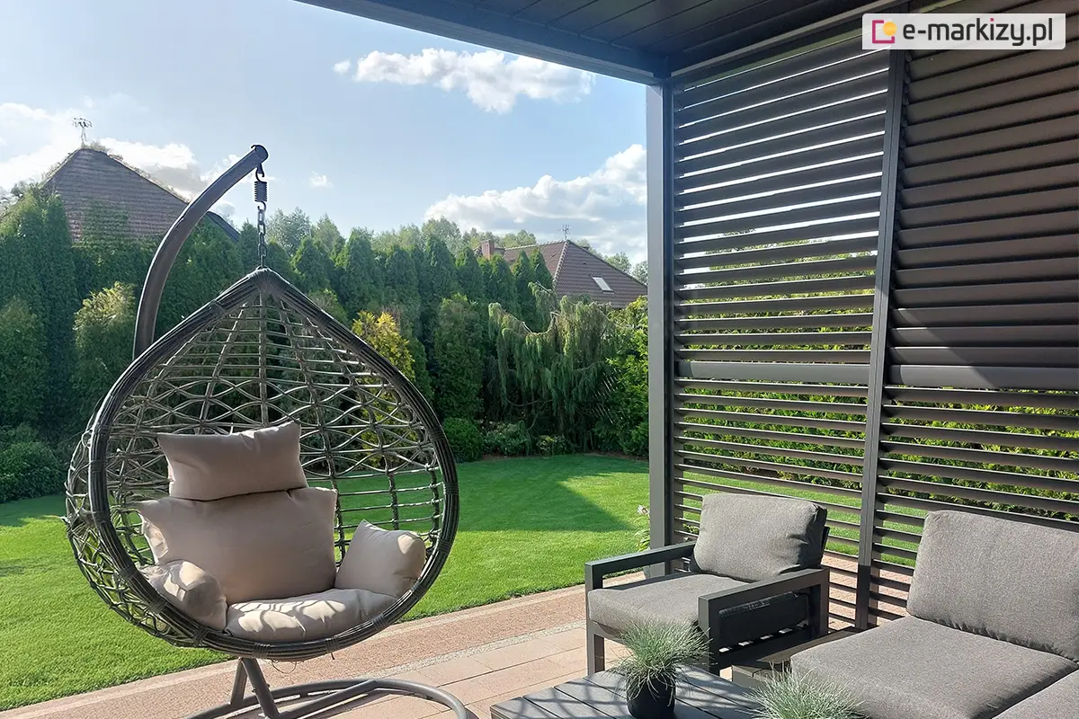 Nowoczesna pergola tarasowa z grafitowymi shuttersami aluminiowymi i szarym fotelem wiszącym typu kokon na tle ogrodu.